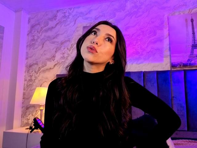 MiaReed with long dark hair, black sweater, and hand on hip, looking up with pouty lips, holding a purple remote, in a room with purple and blue lighting.