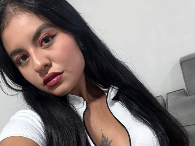 AlessiaVeega with long black hair, red eyeliner, and pink lipstick, wearing a white shirt, showing a small tattoo on her chest, and a slight pout. Her tits are visible.
