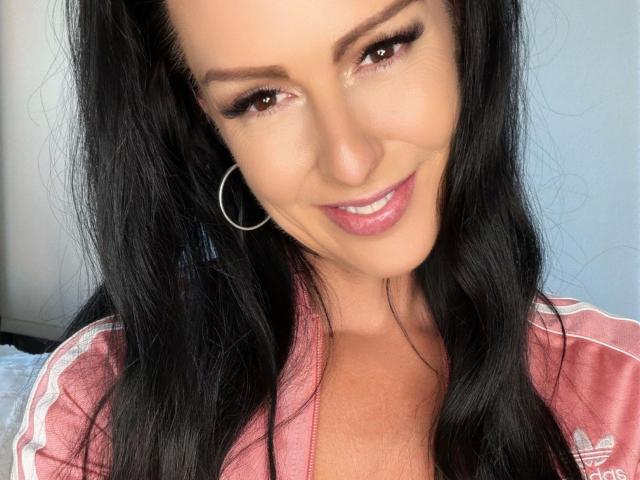 Texaspatti, aantrekkelijk met zwart haar, bruine ogen en roze lippenstift, lacht naar de camera, draagt een roze Adidas trainingsjack met witte strepen en zilveren oorringen..