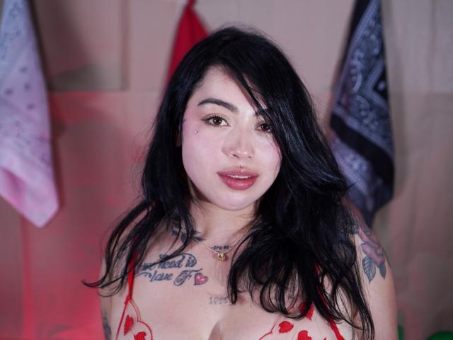 AleStormn, com cabelo preto, batom vermelho, tatuagens e um piercing no nariz, vestindo um sutiã vermelho com bolinhas, olhando para a câmera com a boca ligeiramente aberta. Seios visíveis..