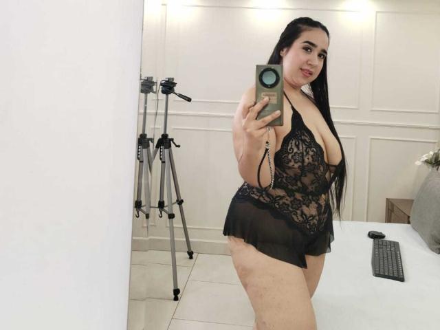 JelenaHunt, met lang zwart haar, draagt zwarte kant lingerie, waarbij haar grote borsten en ronde billen zichtbaar zijn. Ze maakt een selfie met een groene telefoon in een lichte kamer..