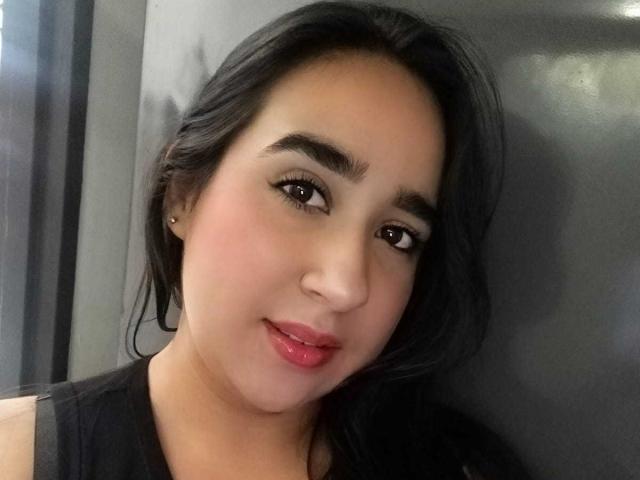 JelenaHunt tiene un rostro adorable con cabello oscuro y ondulado, ojos marrones y lápiz labial rosa. Lleva una blusa negra y tiene un arete pequeño y discreto en la oreja izquierda..