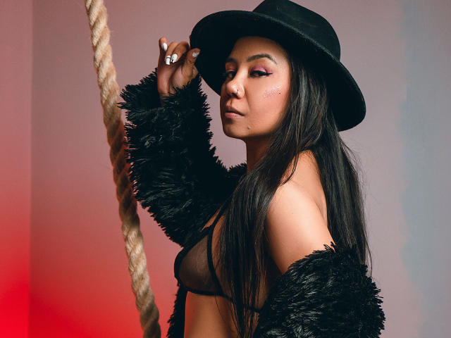 MissCharlotteJolie with long black hair, wearing a black hat and fuzzy jacket, exposing her small tits in a black bra, holding a rope, with pink eyeshadow and a ring.