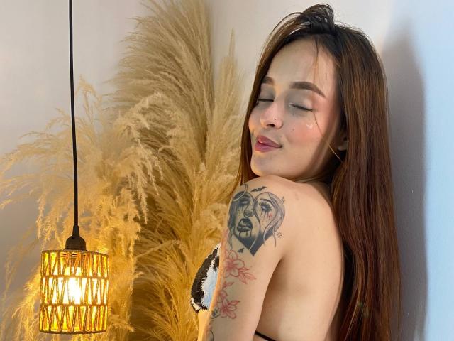 KittyMiller with long brown hair, closed eyes, and a smile, showing tattoos on her arm and side, wearing a black and white bra, under a yellow pampas grass lamp.