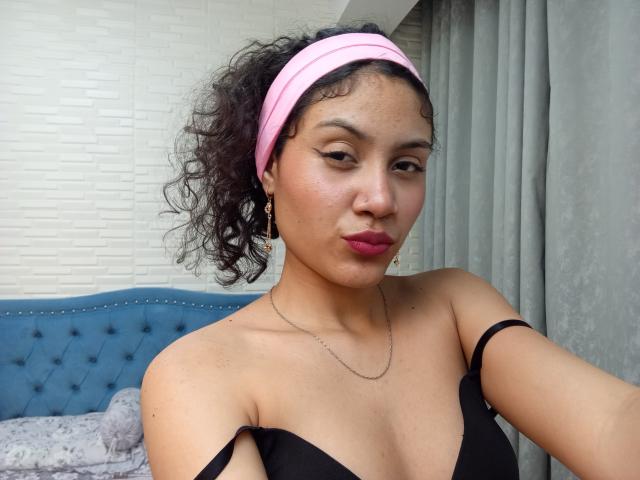 Valery Poleth mit lockigem, schwarzem Haar, einem pinken Stirnband, einem schwarzen Top mit Trägern, kleinen Brüsten, dunkelrosa Lippenstift, goldenen Ohrringen, einer silbernen Halskette und einem schmollenden Gesicht, im Schlafzimmer, blickt in die Kamera..