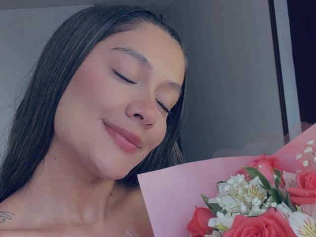 ThaliaAres, les yeux fermés, un sourire doux, tenant un bouquet rose de roses et de fleurs blanches, cheveux foncés, peau claire, maquillage discret, petit tatouage sur l'épaule droite..