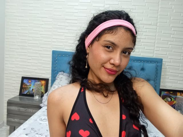 ValeryPoleth, com cabelos pretos encaracolados, uma faixa de cabeça rosa e um top de biquíni vermelho com estampa de coração, sorri para a câmera, mostrando seus seios e batom rosa. Ela tem pele de tom médio e brincos de ouro..