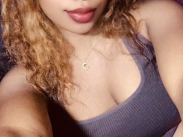TiakoI has curly brown hair, a small gold necklace, and is wearing a grey ribbed tank top with visible cleavage. Her lips are slightly parted.