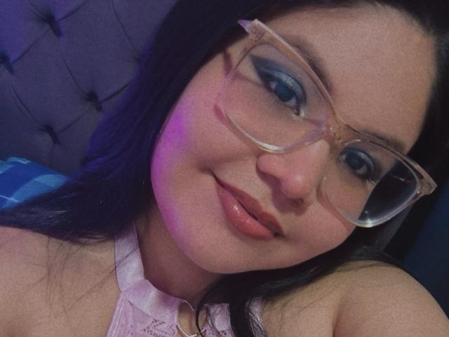 Kappuru with blue eyeshadow, clear glasses, and pink lipstick, smiling slightly, showing her cute nose and glossy lips, wearing a white lace top, lying down.