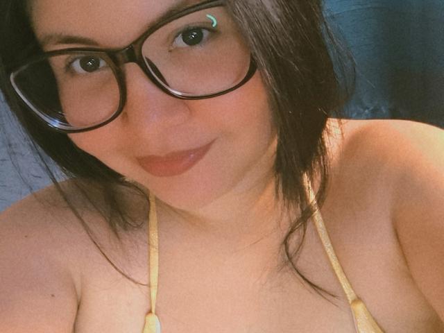 Kappuru with dark glasses, brown hair, and a yellow halter top, showing her tits and a subtle smile. She has a green stud in her left eye.
