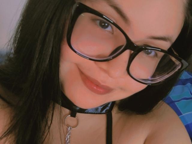 Kappuru with black glasses, wearing a black collar with a ring and chain, smiling at the camera. Her dark hair and red lips are visible. Her tits are partly shown.