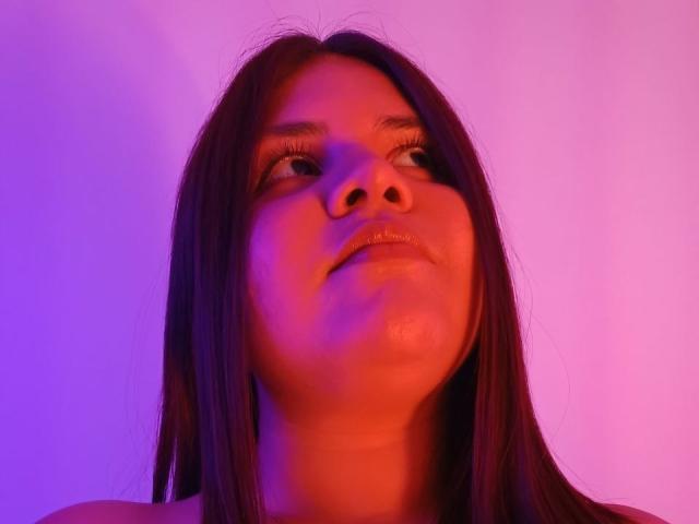 Kappuru has a chubby face with shiny lips, straight brown hair, and is looking up with a slight smile, bathed in purple and red lighting.