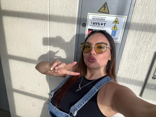 GalaMalkova with brown hair, wearing yellow aviator sunglasses, denim overalls, black tank top, pouting lips, making a peace sign, and a green ring, standing in sunlight.