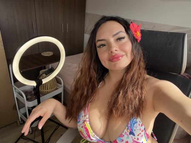 GalaMalkova with medium tits in a colorful floral bikini, ring light on left, black chair behind, pink flower in hair, smiling, holding camera, wavy brown hair, pink lipstick.