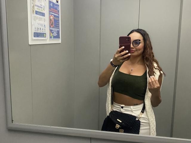 GalaMalkova, con i capelli castano ondulati, occhiali, top verde a vita corta, pantaloni bianchi a vita alta, borsa nera e cardigan bianco aperto, sta scattando un selfie allo specchio in un ascensore..