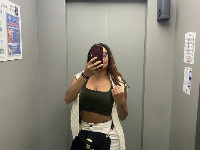 GalaMalkova with tan skin, brown wavy hair, green crop top, white pants, holding phone covering face, white open jacket, black belt, mirror selfie in elevator.