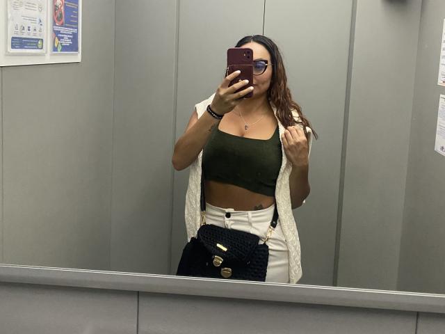 GalaMalkova, con i capelli castani, che indossa occhiali, una canottiera verde, pantaloni bianchi e un gilet bianco, sta scattando un selfie allo specchio in un ascensore, tenendo in mano una borsa nera..
