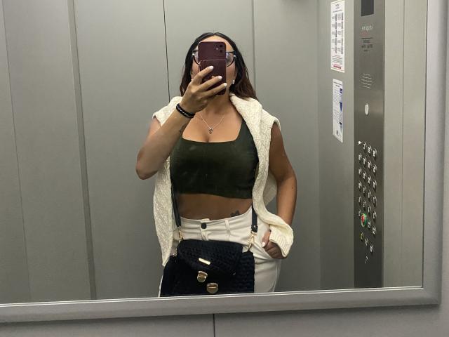 GalaMalkova con capelli castani, top verde, pantaloni bianchi a vita alta e gilet di lana senza maniche, che tiene un telefono, selfie allo specchio nell'ascensore, seno di media grandezza, mano in tasca, braccialetti..