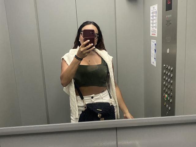 GalaMalkova taking a mirror selfie in an elevator, wearing a green crop top, white open jacket, high-waisted blue jeans, and a dark crossbody bag, holding a red phone.