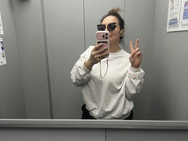 GalaMalkova con gli occhiali, un maglione bianco, pantaloni neri e un telefono rosa, che fa un segno di pace in un selfie allo specchio, con le cuffie nelle orecchie, in un ascensore..