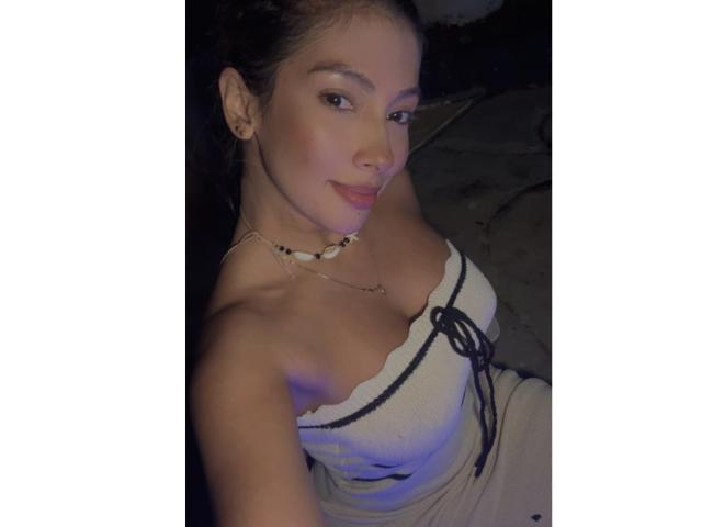 KendraMiler has light brown skin, small nose, and full lips. She's wearing a strapless white top, showing her tits. She's smiling, with a shell necklace, in a dim light.