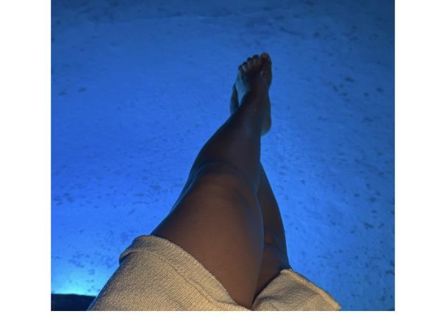 De donkerbruine benen van KendraMiler, gekruist bij de enkels, draagt beige shorts, met haar blote voeten omhoog gericht, tegen een blauw verlichte achtergrond..