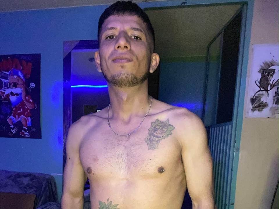 Shirtless AlexBigTool with a tattoo of a rose on his right chest, short dark hair, and a stubble, standing in a room with blue lighting, looking at the camera.