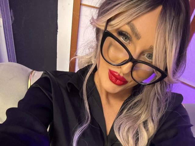 TaraSaint mit blonden, ombre-gefärbten Haaren, roten Lippen, einer schwarzen Brille und einem schwarzen Hemd, die in ihrem Selfie ihr glänzendes Hinterteil zur Schau stellt..