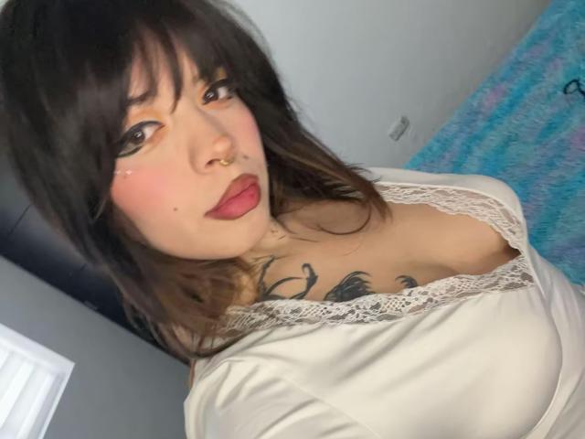 BabeSix with dark brown hair, bangs, nose ring, and pink lipstick, showing cleavage in a lace-trimmed white top, and a tattoo on her chest, pouting slightly.