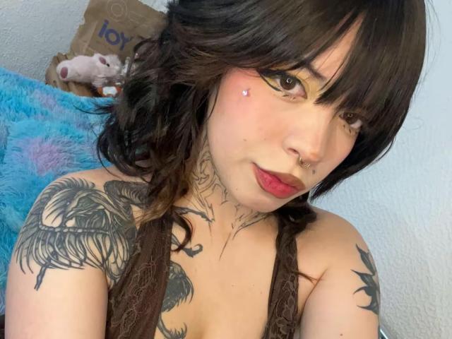A BabeSix tem cabelo escuro e ondulado, olhos castanhos com delineador amarelo, tatuagens nos ombros e no peito, um piercing no nariz e batom vermelho. Ela está sem blusa, mostrando seus seios e tatuagens no pescoço..