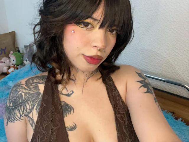 BabeSix met donker, golvend haar, rode lippenstift, een neusring en tatoeages, draagt een bruin kanten topje, waarbij haar decolleté en tatoeages op haar schouders zichtbaar zijn, en zit op een blauw, pluizig oppervlak..