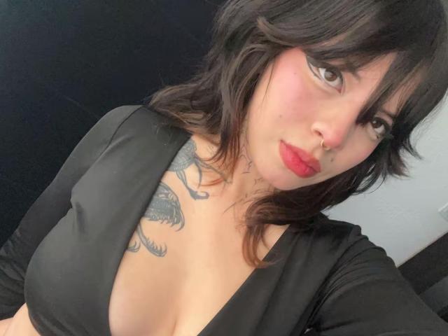 BabeSix, com cabelo preto ondulado, batom vermelho e piercing no nariz, vestindo uma blusa preta que expõe seus seios e uma tatuagem no peito. Ela está com uma expressão sedutora..