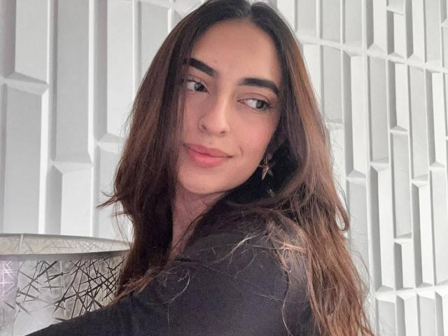 CelesteLarson has long brown hair, full eyebrows, and a slight smile. She's wearing a black top and star earrings, looking over her shoulder. Her tits are subtle.