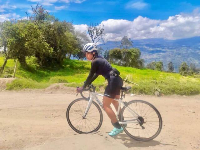 CatalinaReiyna's ass is in tight black cycling shorts, wearing a white helmet, black long-sleeve, and turquoise shoes, riding a white bike on a dirt path.