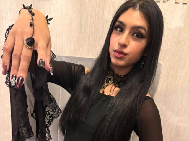 LilithDesire with long black hair, dark eyeliner, and red lipstick, wearing a black lace choker and dress, holding black lace panties with spiked nails and ring.