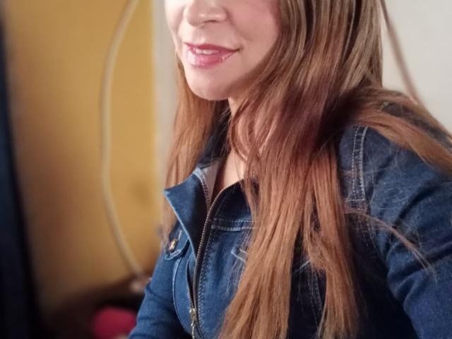 Crisht with long brown hair, blue denim jacket, and pink lipstick, smiling slightly. Her tits are barely visible under the jacket. She's a cute, sexy chick.