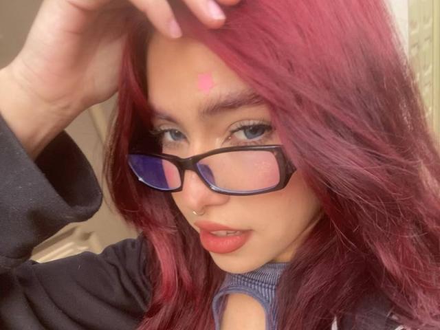 CherryMorganHot, con cabello rojo, gafas negras, piercing en la nariz y lápiz labial rosa, mostrando una blusa con un corte, tocándose la cabeza, con una pequeña marca rosa en la frente..