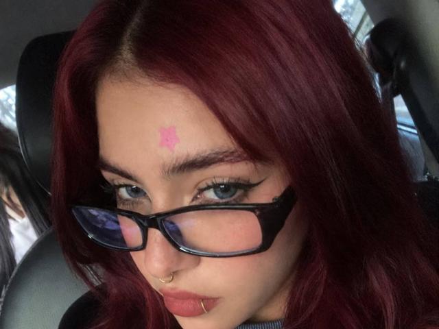 CherryMorganHot, com cabelo ruivo, óculos pretos, piercing no nariz e uma tatuagem de estrela rosa na testa, olhos azuis e lábios rosados, sentada em um carro..