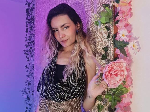 AlanisBlanc with wavy blonde hair, wearing a black and gold sequined top, standing near pink flowers and a large rose, smiling softly, with purple and blue lighting.