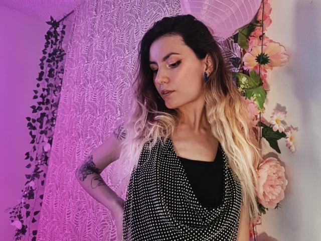 AlanisBlanc with long blonde hair, black nose ring, and tattoos, wearing a black and white polka dot dress, standing with eyes closed, in a pink-lit room.
