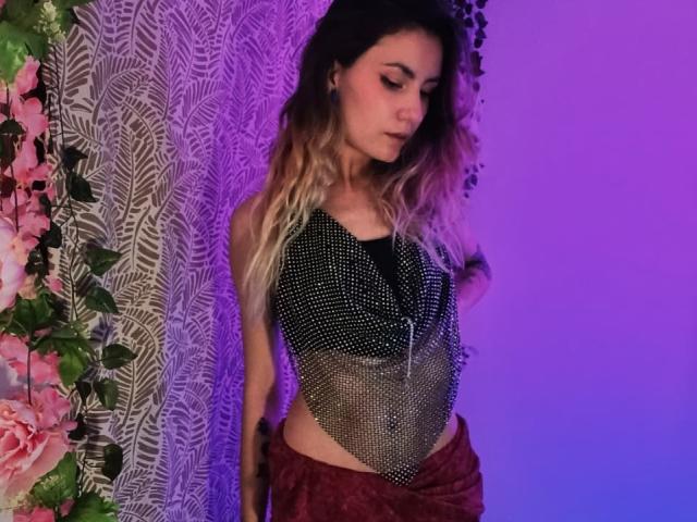 AlanisBlanc with long blonde hair, wearing a sparkly black top and red pants, standing against a patterned curtain with pink flowers, in purple and blue lighting.