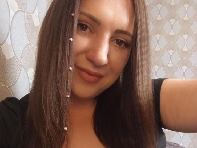 TinaEves hat braunes Haar, einen Nasenring und ein leichtes Lächeln. Sie trägt ein schwarzes Oberteil mit geflochtenem Haar, das mit weißen Perlen verziert ist. Ihre Brüste sind klein..