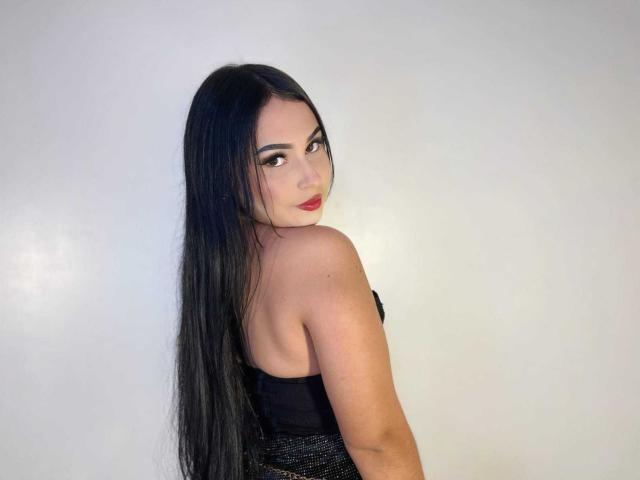 HannahBecker with long black hair, looking over her shoulder, wearing a strapless black dress, showing her bare back and red lips, standing against a plain white wall.