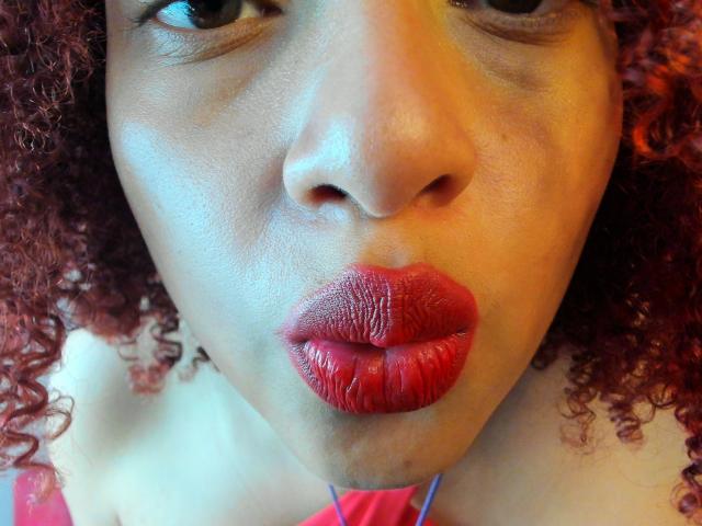 Het gezicht van VioletaSwitch, met krullend rood haar, donkerbruine huid en glanzende rode lippen, met haar volle lippen samengeknepen. Haar ogen zijn lichtjes samengeknepen, en haar neus is gecentreerd..
