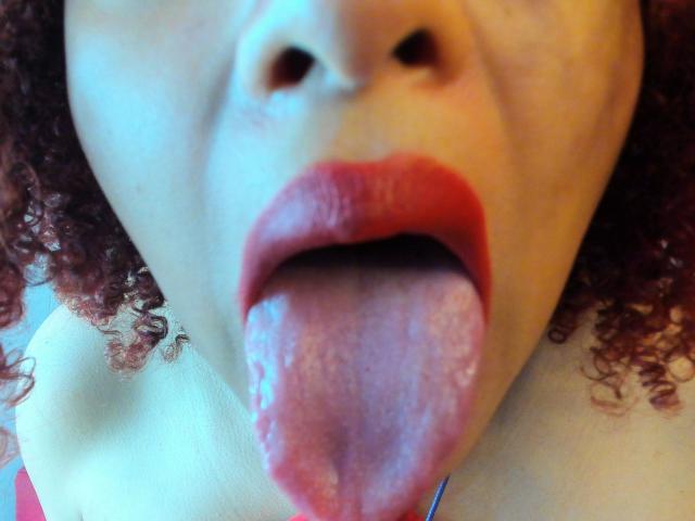 Le visage de VioletaSwitch, avec ses cheveux roux bouclés, sa peau pâle et ses lèvres rouges. Sa langue est dehors, humide et rose, recouvrant sa lèvre inférieure. Son nez est centré en haut..