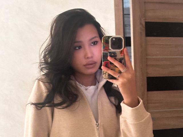 LeonilaSeewald with wavy black hair, wearing a beige zip-up hoodie, holding a phone with a colorful case, taking a mirror selfie, showing a calm, slightly pouty expression.