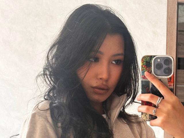 LeonilaSeewald with long black wavy hair, wearing a beige hoodie, taking a selfie with a colorful phone case, showing a slight pout, and a small beauty mark on her nose.