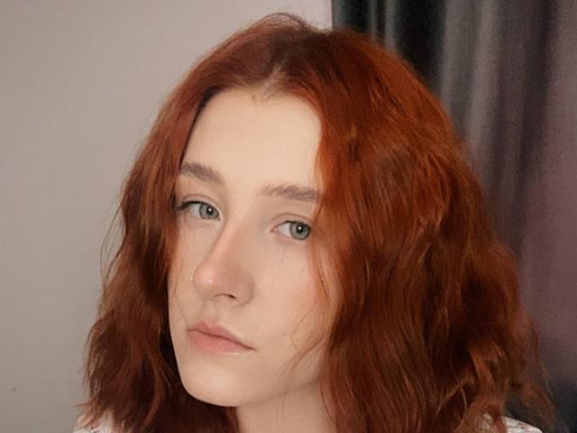 MalvinaHauenstein has wavy red hair, green eyes, and a pale complexion with slightly pouty, pinkish lips. Her nose is small and she looks neutral.