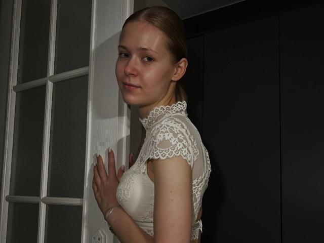 LillianaWhang with light skin and brown hair, wearing a white lace top, standing by a door, hands on it, looking over her shoulder with a slight smile.