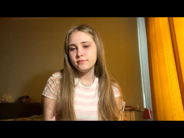 KaleighLopera with long blonde hair, wearing a pink and white striped shirt, has a bored or slightly annoyed expression, sitting in a yellow-walled room with orange curtains.